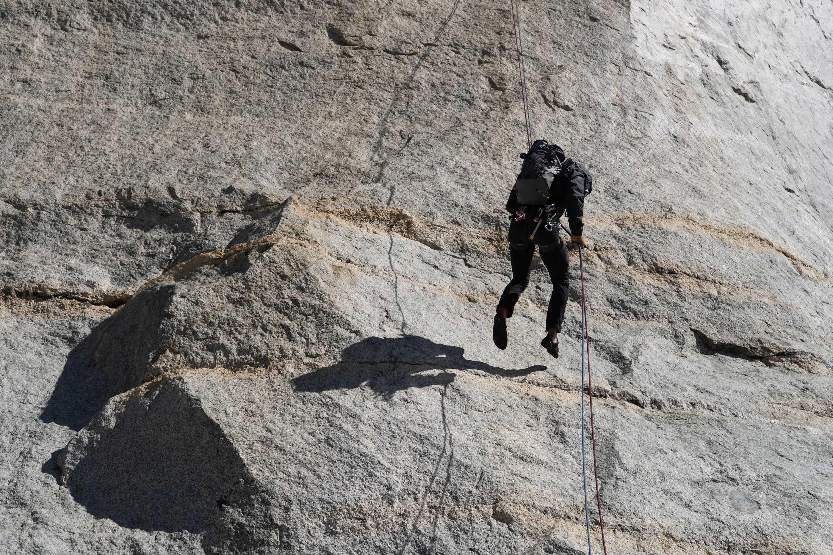 A climber rappelling down a sunlit granite slab, a sharp shadow on the rock below.